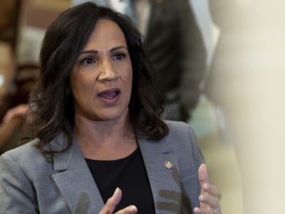 An image of Minnesota House Speaker Lisa Demuth, a woman with shoulder-length, dark wavy hair speaks and gestures with both hands while wearing a gray blazer over a black top.