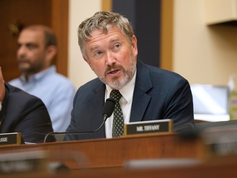 U.S. Representative Thomas Massie, a man with wavy gray hair and a beard wears a navy blue suit jacket and a patterned tie as he speaks into a microphone at a Congressional hearing.