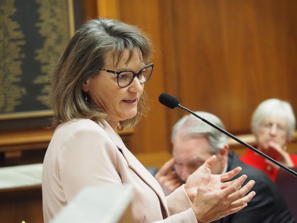 Indiana state Representative Wendy Dant Chesser, a woman in a pink blazer and glasses speaks into a microphone, gesturing with her hands. Two people listen in the background.