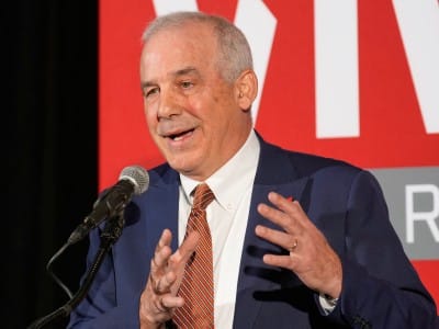 Ohio House Speaker Matt Huffman speaks into a microphone while gesturing with both hands. He wears a blue suit jacket, a white shirt, and an orange and white striped tie. The background features a bright red backdrop with partial large white letters and a grey banner displaying the letter "R".