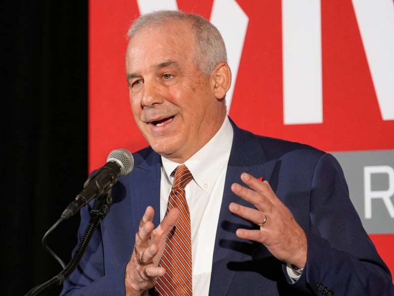 Ohio House Speaker Matt Huffman speaks into a microphone while gesturing with both hands. He wears a blue suit jacket, a white shirt, and an orange and white striped tie. The background features a bright red backdrop with partial large white letters and a grey banner displaying the letter "R".