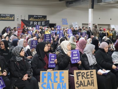 A large indoor crowd, including many women wearing hijabs, is gathered for a community rally or demonstration. Attendees hold small purple signs and large cardboard placards with the text "FREE SALAH" written in blue and black. In the background, black banners with gold lettering read "Bismillah" and "Alhamdulillah" next to a sign that says "FREE."