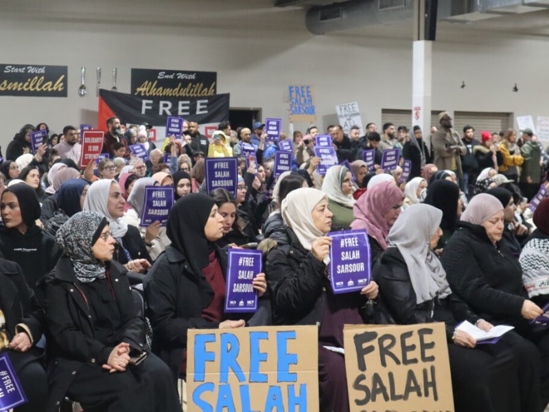 A large indoor crowd, including many women wearing hijabs, is gathered for a community rally or demonstration. Attendees hold small purple signs and large cardboard placards with the text "FREE SALAH" written in blue and black. In the background, black banners with gold lettering read "Bismillah" and "Alhamdulillah" next to a sign that says "FREE."