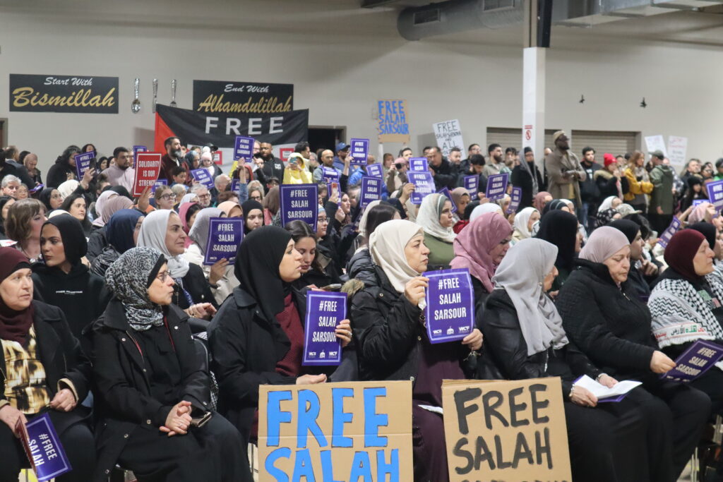 A large indoor crowd, including many women wearing hijabs, is gathered for a community rally or demonstration. Attendees hold small purple signs and large cardboard placards with the text "FREE SALAH" written in blue and black. In the background, black banners with gold lettering read "Bismillah" and "Alhamdulillah" next to a sign that says "FREE."