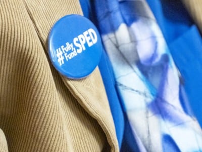 A close-up photograph focuses on a bright blue circular pin attached to the lapel of a tan corduroy jacket. The pin features white text that reads "#Fully Fund SPED." In the softly blurred background, a vibrant blue shirt and a patterned purple-and-blue scarf are visible