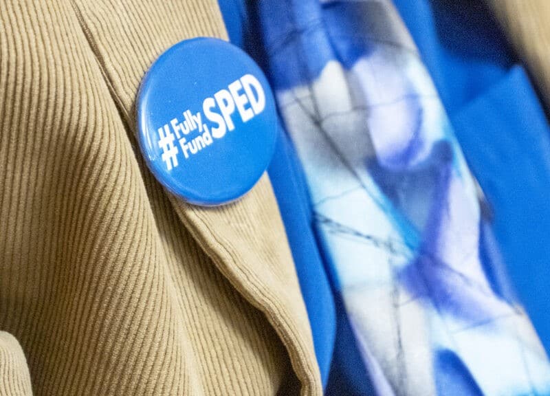 A close-up photograph focuses on a bright blue circular pin attached to the lapel of a tan corduroy jacket. The pin features white text that reads "#Fully Fund SPED." In the softly blurred background, a vibrant blue shirt and a patterned purple-and-blue scarf are visible