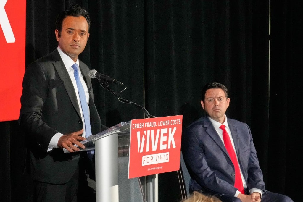 Vivek Ramaswamy stands at a podium speaking, wearing a dark suit and light blue tie against a black backdrop. A bright red sign on the podium reads, "CRUSH FRAUD. LOWER COSTS. VIVEK FOR OHIO" in white text. Seated to the right, his running mate Rob McColley in a dark suit and a vibrant red tie watches the presentation.