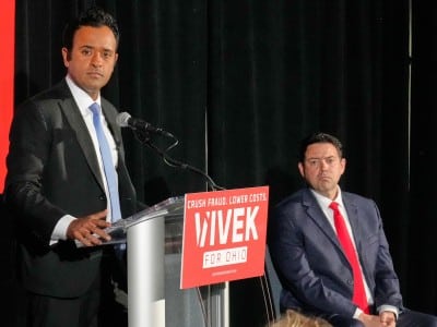 Vivek Ramaswamy stands at a podium speaking, wearing a dark suit and light blue tie against a black backdrop. A bright red sign on the podium reads, "CRUSH FRAUD. LOWER COSTS. VIVEK FOR OHIO" in white text. Seated to the right, his running mate Rob McColley in a dark suit and a vibrant red tie watches the presentation.