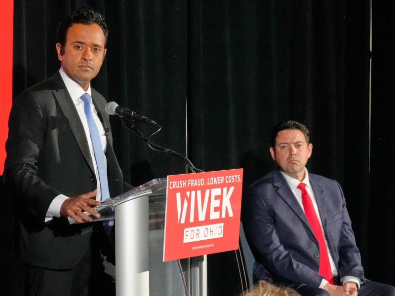 Vivek Ramaswamy stands at a podium speaking, wearing a dark suit and light blue tie against a black backdrop. A bright red sign on the podium reads, "CRUSH FRAUD. LOWER COSTS. VIVEK FOR OHIO" in white text. Seated to the right, his running mate Rob McColley in a dark suit and a vibrant red tie watches the presentation.