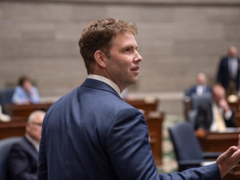 Missouri state Senator Stephen Webber, a man with wavy brown hair wearing a blue suit jacket gestures with his hand while speaking in a legislative chamber. He is captured from a three-quarters back view, looking upward and to the right.