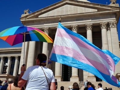 A person viewed from behind holds a vibrant rainbow umbrella alongside a large, waving transgender pride flag with light blue, pink, and white stripes. They are standing with a crowd in front of a grand stone building featuring classical columns and the engraved text "STATE OF OKLAHOMA."