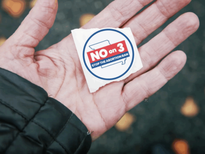 A closeup shot of a hand holding a voting sticker that says, "NO on 3. Stop the abortion ban."