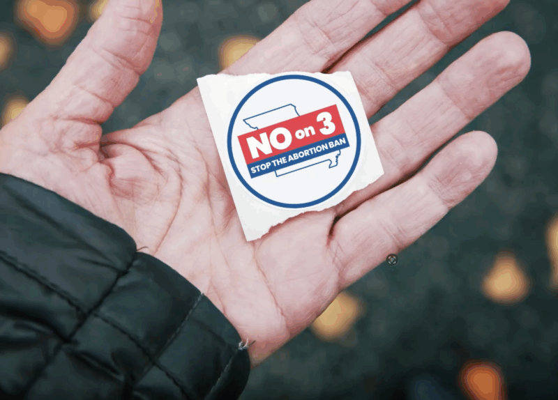 A closeup shot of a hand holding a voting sticker that says, "NO on 3. Stop the abortion ban."