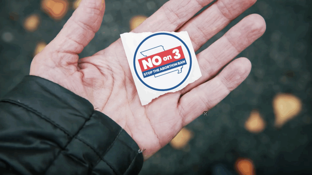A closeup shot of a hand holding a voting sticker that says, "NO on 3. Stop the abortion ban."
