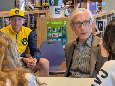 Wisconsin Governor Tony Evers, wearing glasses and a grey suit jacket, speaks with a group of students inside a library. In the background, a boy sits wearing a yellow and navy Milwaukee Brewers hat and jacket near a green poster that reads "WISCONSIN".