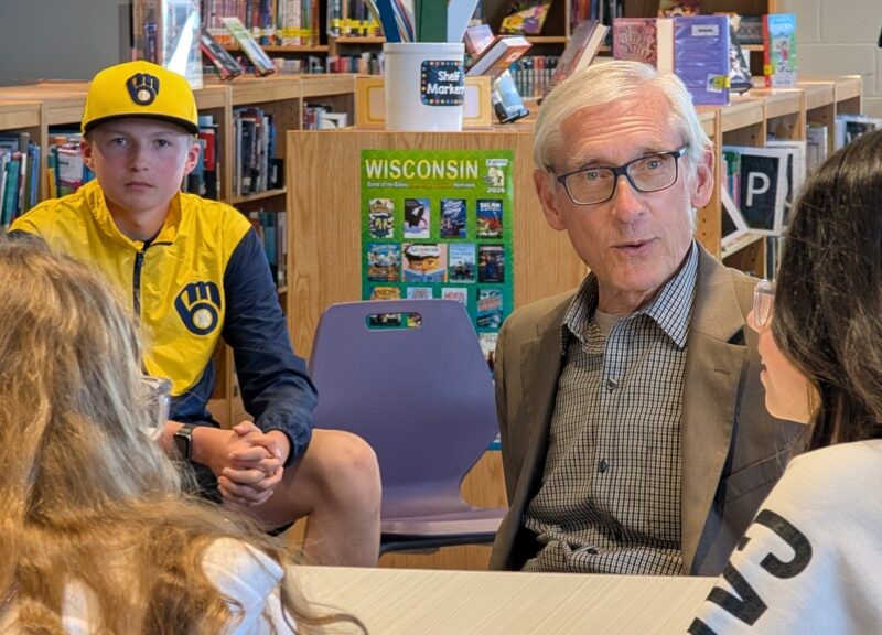 Wisconsin Governor Tony Evers, wearing glasses and a grey suit jacket, speaks with a group of students inside a library. In the background, a boy sits wearing a yellow and navy Milwaukee Brewers hat and jacket near a green poster that reads "WISCONSIN".