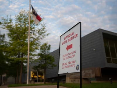 A white sign in the foreground features a red left-pointing arrow and text that reads "OFFICIAL VOTE CENTER" in English, Spanish, and Vietnamese. In the background, a Texas state flag flies on a flagpole outside a modern grey building surrounded by green trees. The scene is set outdoors under a bright, cloudy sky.