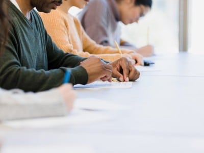 A row of people sit at a long white table focused on writing. In the center, a man in a green sweater writes on a paper with a pencil. Other individuals are softly blurred in the background.