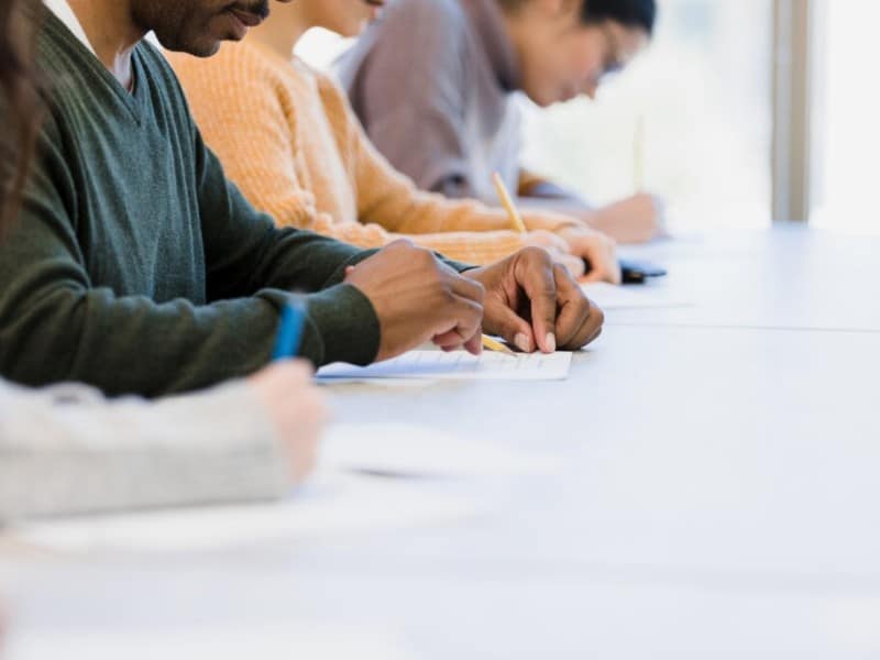 A row of people sit at a long white table focused on writing. In the center, a man in a green sweater writes on a paper with a pencil. Other individuals are softly blurred in the background.