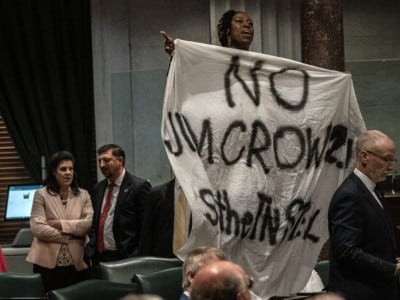 Tennessee state Senator Charlane Oliver stands in a legislative chamber holding up a large white banner with spray-painted text that reads "NO JIM CROW 2.0!" while pointing upward.