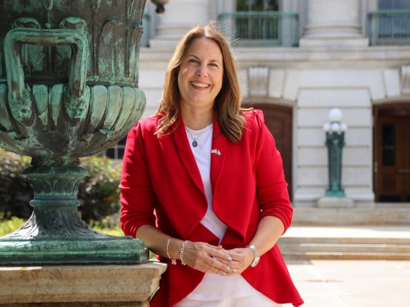 Wisconsin state Senator Dianne Hesselbein, smiling with shoulder-length brown hair, poses outdoors in front of the Wisconsin State Capitol a blurred background. She is leaning against a large, ornate green and black copper sculpture on a stone pedestal with her hands clasped in front of her. She is wearing a vibrant red open blazer over a white top and white pants, accessorized with a pendant necklace, a small lapel pin, a watch and bracelets.