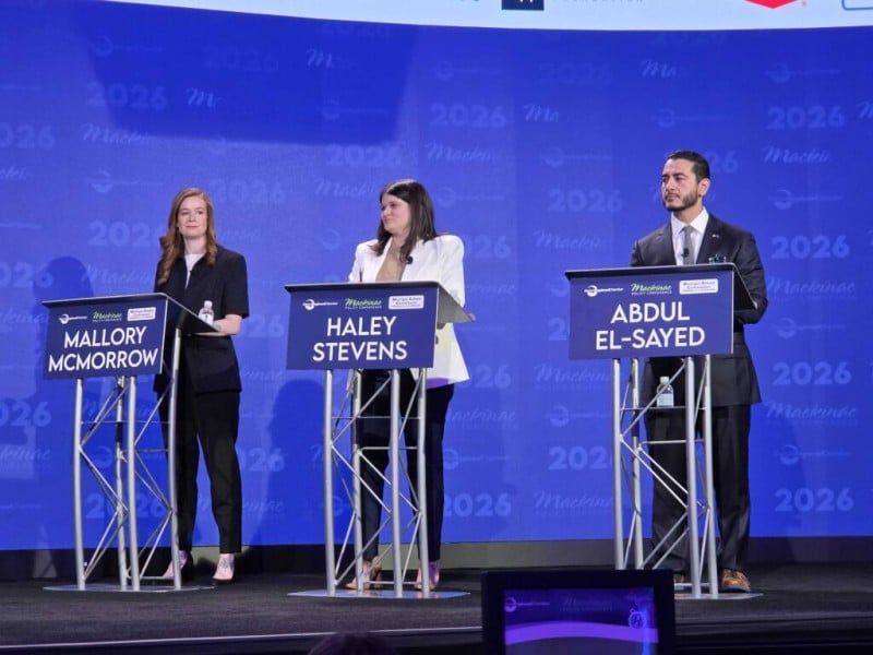 Mallory McMorrow, Haley Stevens, and Abdul El-Sayed stand behind individual metal lecterns with blue nameplates on a stage. The bright blue backdrop features repeating text and logos for the "2026 Mackinac Policy Conference."