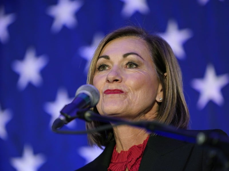 A close-up of Iowa Governor Kim Reynolds, a woman with shoulder-length brown hair looking slightly upward while standing at a microphone. She wears a dark blazer over a vibrant, ruffled red shirt. The background consists of a softly blurred blue backdrop filled with white stars.