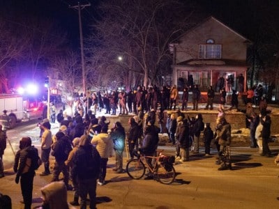A large crowd gathers at night on a street next to a Minneapolis Fire truck with bright, flashing red and blue emergency lights. People stand on the road, sidewalk, and the porch of a two-story house, with several holding up phones to film.