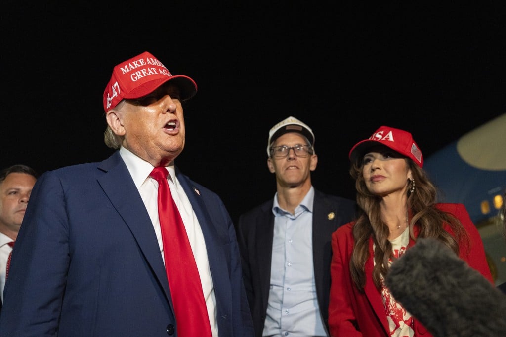Donald Trump speaks outdoors at night wearing a navy blue suit, a bright red tie, and a red hat with the text "MAKE AMERICA GREAT AGAIN." To his right stands Iowa governor candidate Randy Feenstra and former Homeland Security Secretary Kristi Noem.