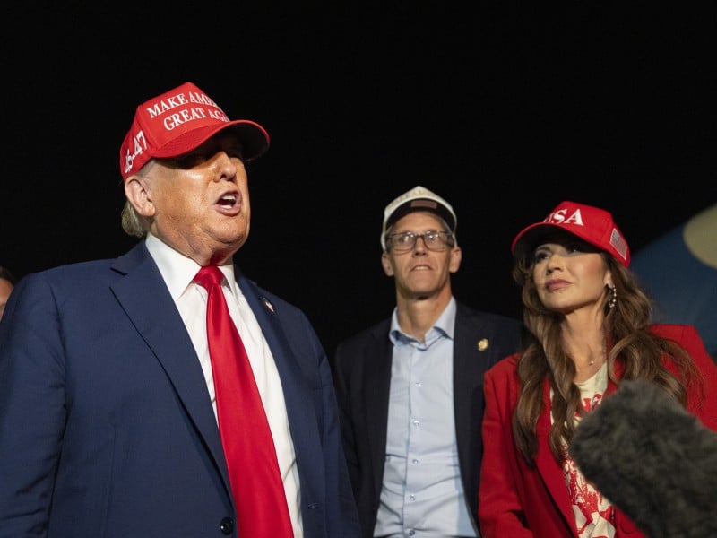 Donald Trump speaks outdoors at night wearing a navy blue suit, a bright red tie, and a red hat with the text "MAKE AMERICA GREAT AGAIN." To his right stands Iowa governor candidate Randy Feenstra and former Homeland Security Secretary Kristi Noem.