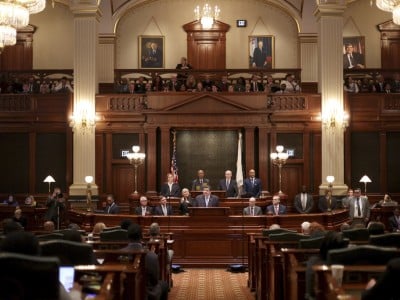 Illinois Gov. JB Pritzker delivers an address from a central wooden podium inside a grand, multi-tiered legislative chamber filled with public officials and audience members. He is dressed in a dark blue suit, a light blue collared shirt and a solid blue necktie, matching the formal business attire worn by the surrounding lawmakers.