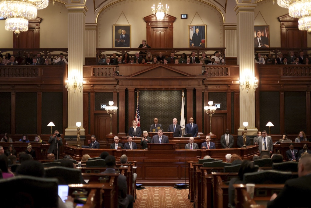 Illinois Gov. JB Pritzker delivers an address from a central wooden podium inside a grand, multi-tiered legislative chamber filled with public officials and audience members. He is dressed in a dark blue suit, a light blue collared shirt and a solid blue necktie, matching the formal business attire worn by the surrounding lawmakers.
