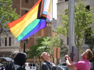 Lansing Mayor Andy Schor and Councilmember Ryan Kost raise a Progress Pride flag on a tall flagpole over the Lansing City Hall.