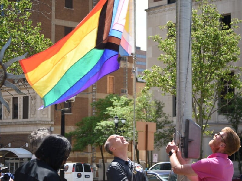 Lansing Mayor Andy Schor and Councilmember Ryan Kost raise a Progress Pride flag on a tall flagpole over the Lansing City Hall.