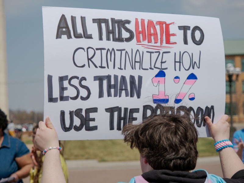 a person seen from behind holds up a large white protest sign outdoors. The handwritten message reads, "ALL THIS HATE TO CRIMINALIZE HOW LESS THAN 1% USE THE BATHROOM," with the word "HATE" in red and the "1%" filled with light blue, pink, and white stripes.