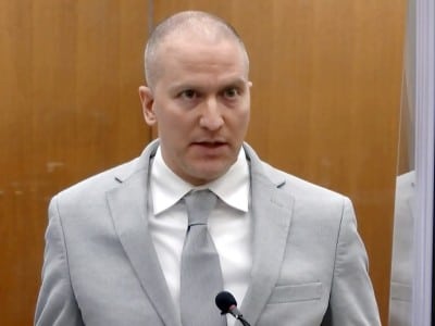 A medium close-up shot shows Derek Chauvin speaking into a microphone with a wide-eyed expression in front of a wood-paneled wall. He is wearing a light gray suit jacket, a white collared shirt and a matching light gray tie.