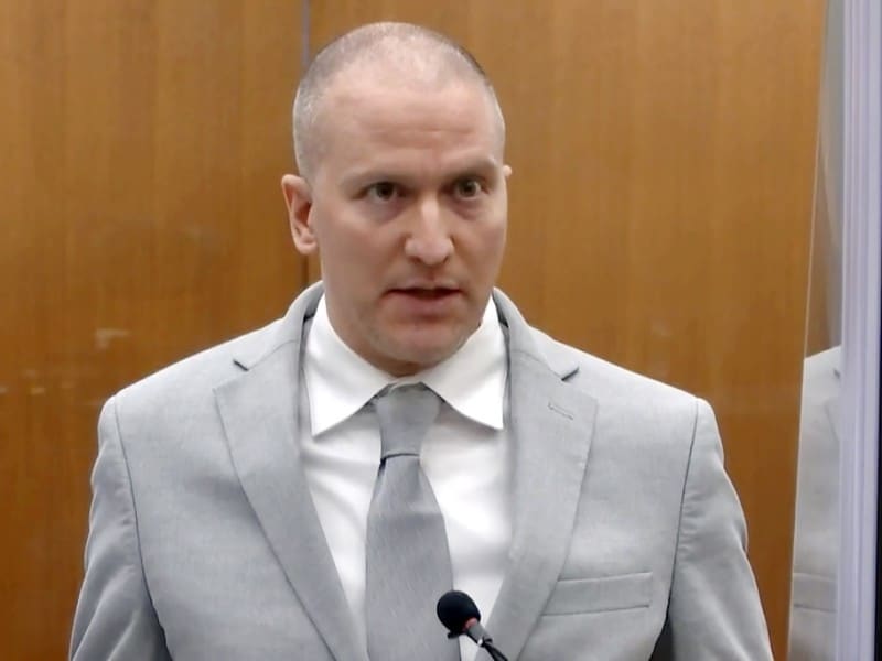 A medium close-up shot shows Derek Chauvin speaking into a microphone with a wide-eyed expression in front of a wood-paneled wall. He is wearing a light gray suit jacket, a white collared shirt and a matching light gray tie.