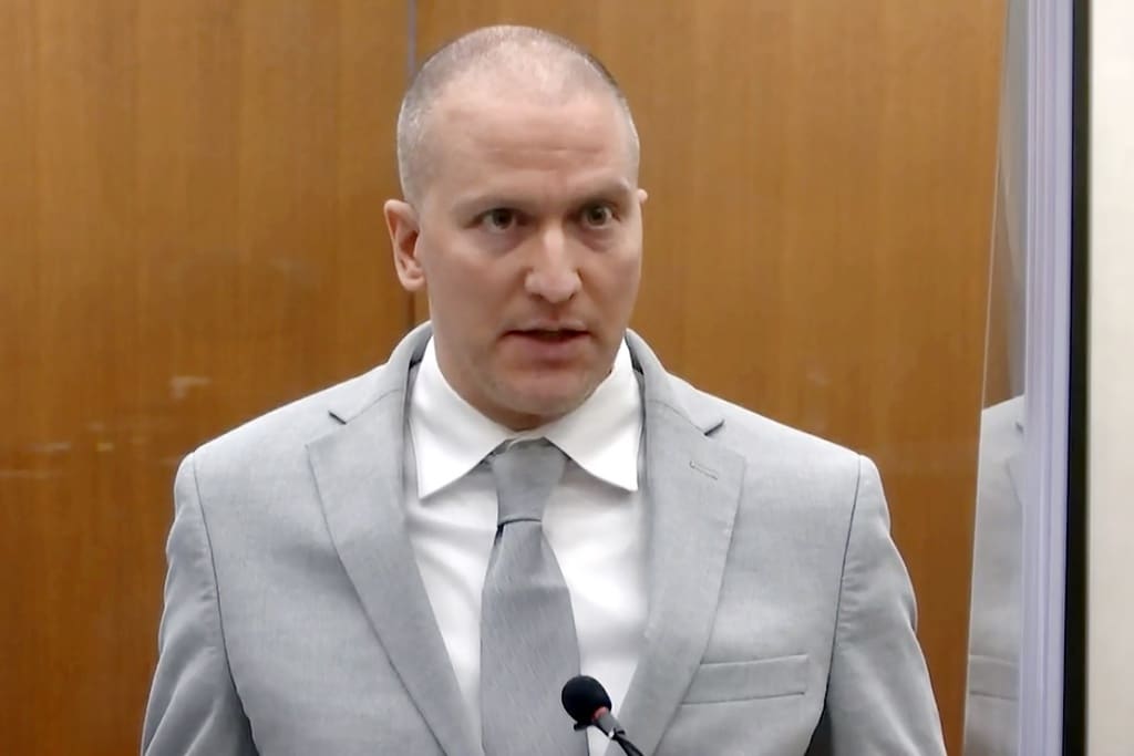 A medium close-up shot shows Derek Chauvin speaking into a microphone with a wide-eyed expression in front of a wood-paneled wall. He is wearing a light gray suit jacket, a white collared shirt and a matching light gray tie.