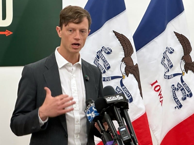A man is speaking at a press conference microphone setup, with his hand raised in mid-gesture and two Iowa state flags draped behind him. He is wearing a dark grey suit jacket over an unbuttoned, collared white dress shirt.
