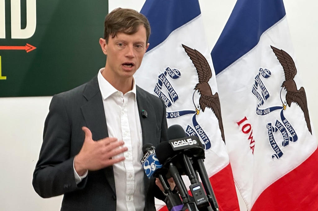 A man is speaking at a press conference microphone setup, with his hand raised in mid-gesture and two Iowa state flags draped behind him. He is wearing a dark grey suit jacket over an unbuttoned, collared white dress shirt.