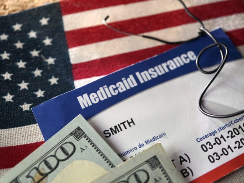 A blue and white Medicaid Insurance card rests on an American flag. A hundred-dollar bill and a pair of eyeglasses lie on top of the card. The document displays the name "SMITH" and coverage dates from 2016.