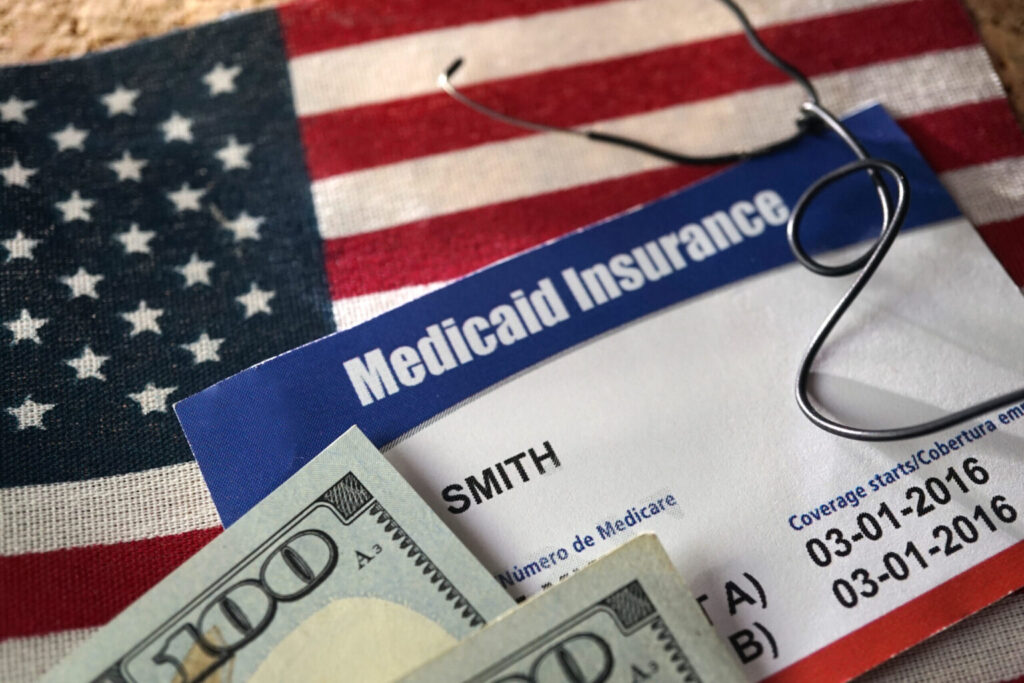 A blue and white Medicaid Insurance card rests on an American flag. A hundred-dollar bill and a pair of eyeglasses lie on top of the card. The document displays the name "SMITH" and coverage dates from 2016.