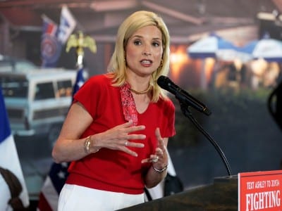 U.S. Rep. Ashley Hinson speaks at a podium wearing a bright red top and white pants. She gestures with her hands in front of flags and a blurred backdrop.