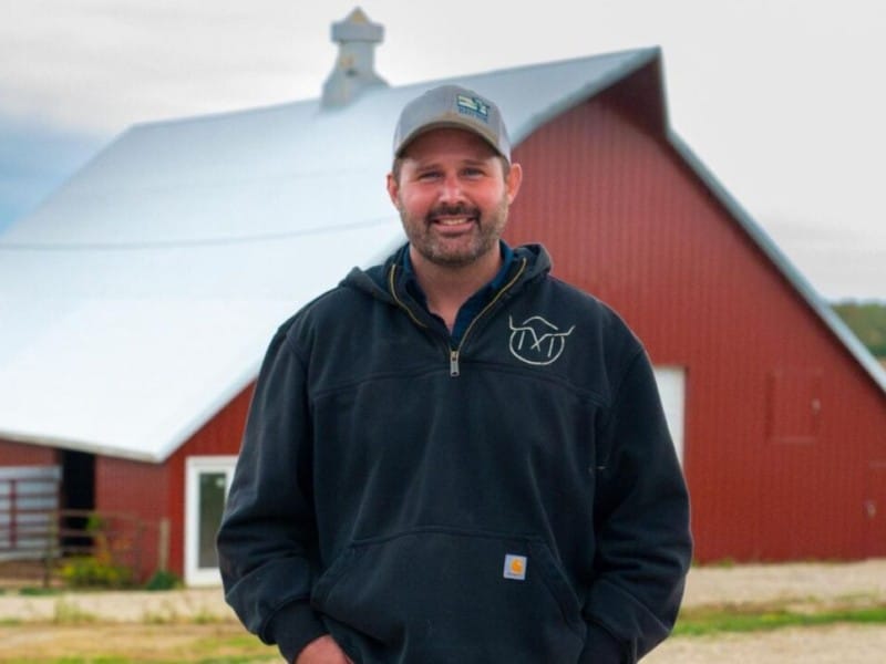 Dave Muhlbauer stands smiling in front of a large red barn with a bright white roof. He wears a grey baseball cap and a black hooded sweatshirt featuring a white steer graphic on the chest.