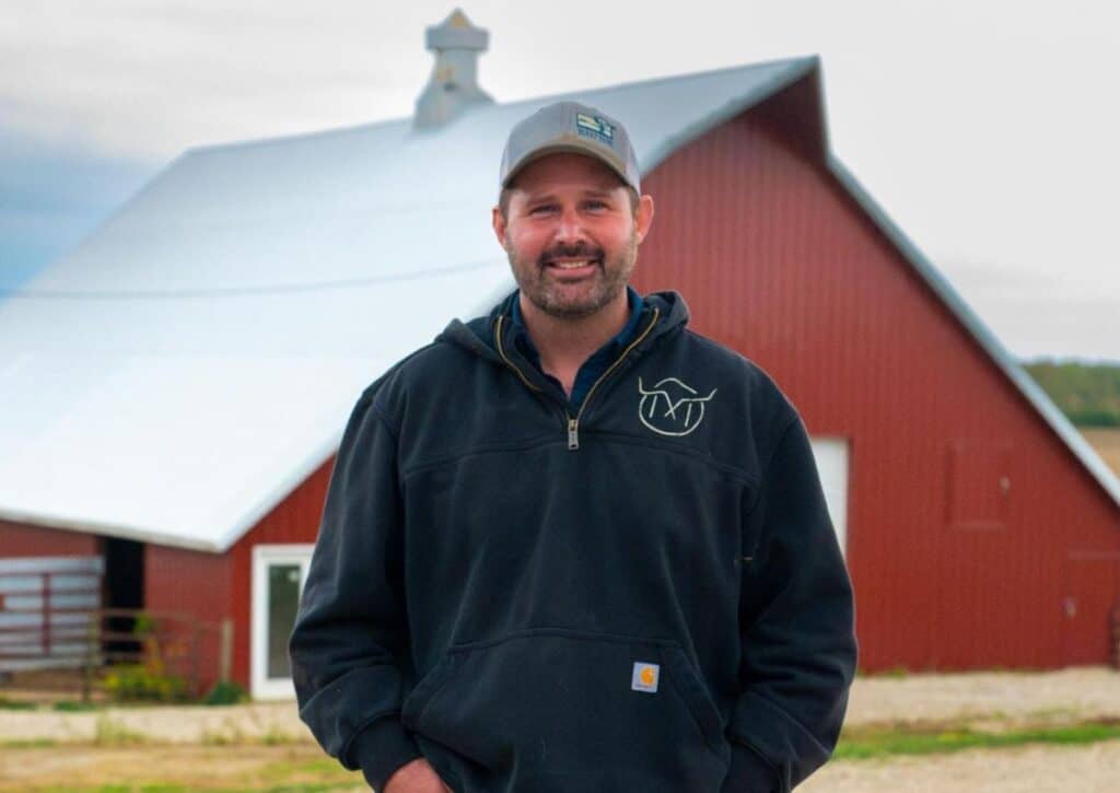Dave Muhlbauer stands smiling in front of a large red barn with a bright white roof. He wears a grey baseball cap and a black hooded sweatshirt featuring a white steer graphic on the chest.