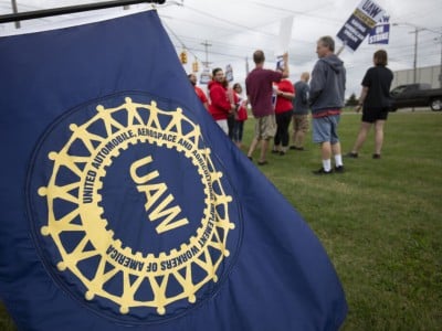 A group of United Auto Workers members striking at the General Motors Lansing plant in 2023. A blue and gold UAW flag appears in the foreground.