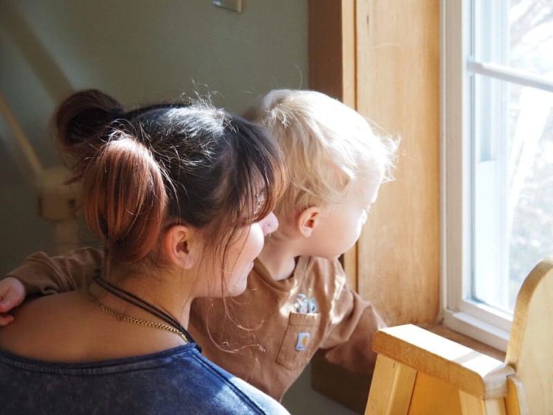 A woman with dark hair styled in buns and a blonde toddler look out a brightly lit window together. Warm, natural sunlight streams into the scene through the wooden window frame.