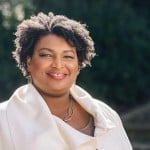 A close-up portrait shows Stacey Abrams smiling warmly outdoors against a blurred background of green foliage. She is wearing a cream-colored jacket or wrap over a brown top, paired with a gold link necklace and matching drop earrings.