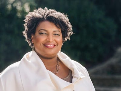A close-up portrait shows Stacey Abrams smiling warmly outdoors against a blurred background of green foliage. She is wearing a cream-colored jacket or wrap over a brown top, paired with a gold link necklace and matching drop earrings.