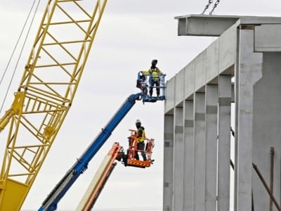 A gray concrete building framework under construction against an overcast sky. On the left, a large yellow crane is visible alongside two workers in high-visibility vests operating blue and orange boom lifts.